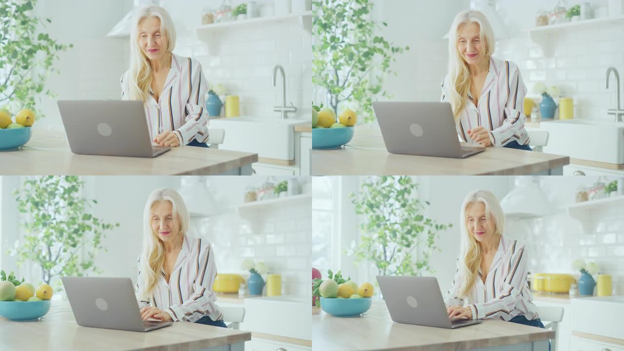 美丽的高级女人使用笔记本电脑，在阳光明媚的厨房里喝一杯咖啡或茶。祖母通过互联网和社交媒体与孩子建立联