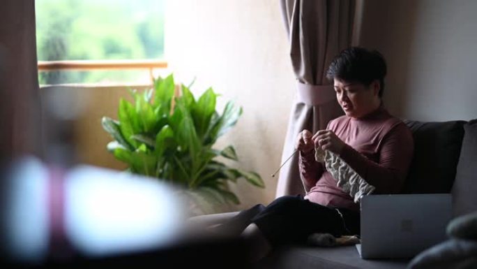 一名亚洲中国中年妇女在客厅休闲活动中编织，并使用笔记本电脑从互联网上学习