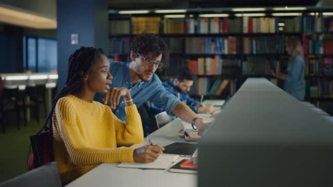 大学图书馆: 有天赋的黑人女孩使用笔记本电脑，聪明的同学解释并帮助她完成课堂作业。快乐多样的学生一起