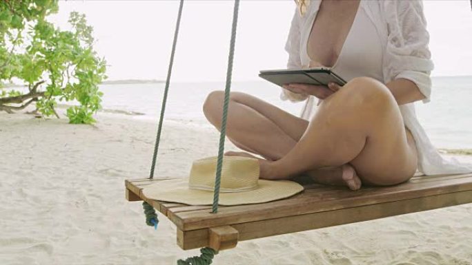 带着数字平板电脑的女士在马尔代夫热带海洋海滩的秋千上放松