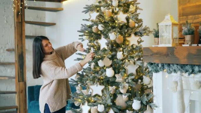 微笑的女孩正在用球和灯光装饰圣诞树，享受假期和创意活动。漂亮的带壁炉的装饰房间是可见的。