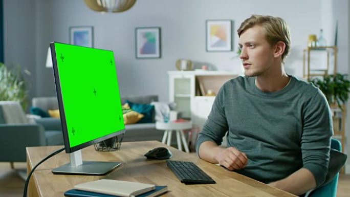 英俊的年轻人坐在舒适的公寓里的办公桌旁，在绿色的模拟屏幕个人电脑上工作。