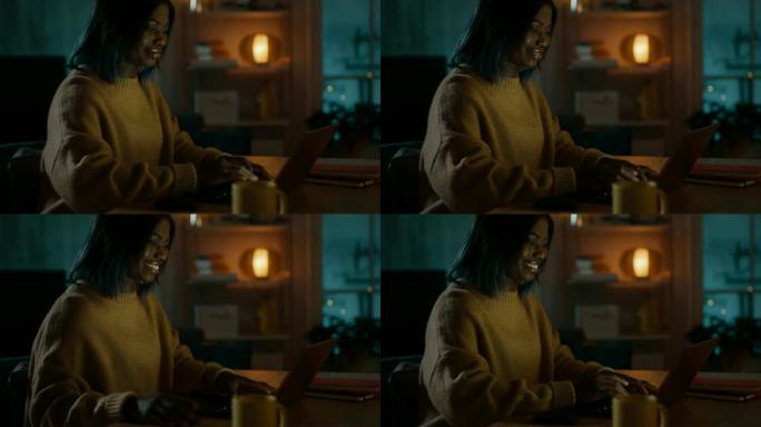 美丽的黑人女孩的肖像坐在家里的办公桌旁使用电脑，她穿着保暖的毛衣。晚上，有创意的女人在舒适的客厅里用