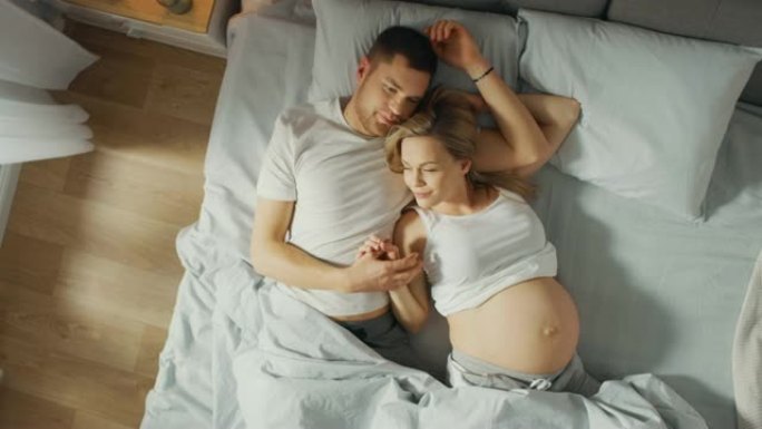 幸福的年轻夫妇在床上拥抱在一起，年轻的女人怀孕了，有爱心的伴侣温柔地抚摸着她的肚子。阳光普照。自上而