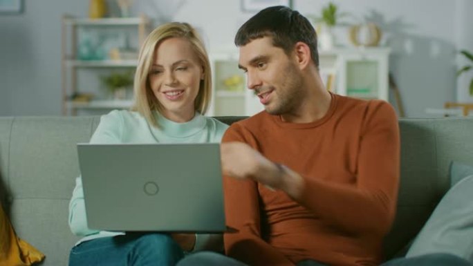可爱的年轻夫妇坐在家里的沙发上，他们使用笔记本电脑，玩得很开心。美丽幸福的年轻夫妇恋爱的肖像。