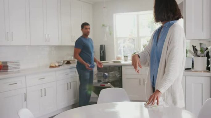 千禧一代妇女清洁厨房桌子，她的伴侣在背景中填充洗碗机