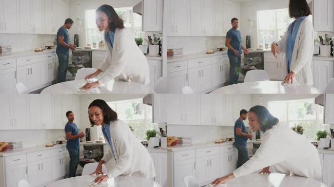 千禧一代妇女清洁厨房桌子，她的伴侣在背景中填充洗碗机