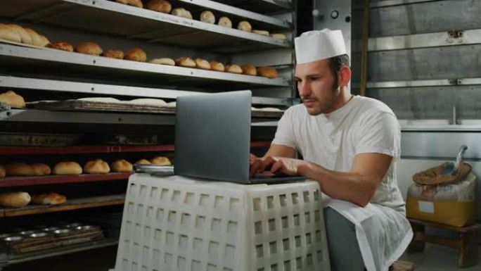 英俊的年轻面包师学生的慢动作使用笔记本电脑进行在线学习，并在面包店学习课程，背景是面包和烤箱。