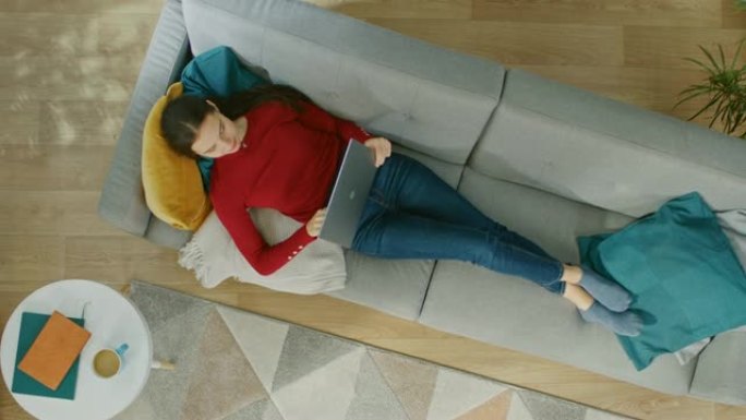 穿着红色上衣和蓝色牛仔裤的年轻女孩躺在沙发上，用笔记本电脑。她抬头微笑。舒适的客厅，配有现代室内植物