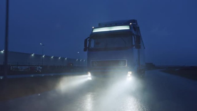 蓝色长途半卡车，满载货物的拖车在夜间在高速公路上行驶，穿过雨、雾、雪穿越大陆。工业仓库区。正面跟随镜