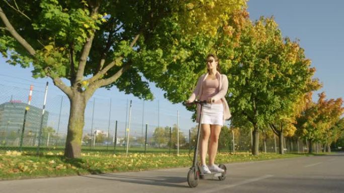 低角度: 快乐的女孩骑着电动滑板车在路上跑过秋天的彩色树木。