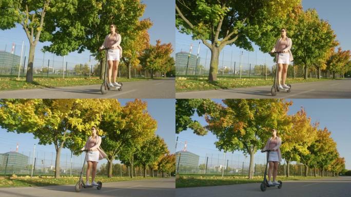 低角度: 快乐的女孩骑着电动滑板车在路上跑过秋天的彩色树木。