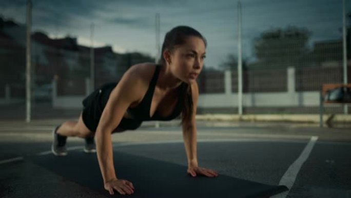 美丽精力充沛的健身女孩做俯卧撑运动。她正在一个有围栏的室外篮球场里锻炼身体。居民区下雨后的晚间录像。