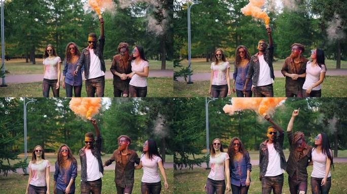 快乐的朋友慢动作，脸上画着脸，拿着烟团在公园里散步，微笑着，明亮的橙色烟雾升起。乐趣和人的概念。