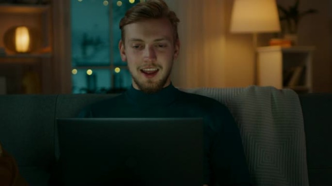 一个英俊的年轻人坐在沙发上的肖像，用笔记本电脑打视频电话，打招呼，挥手致意。晚上，男子使用计算机网络