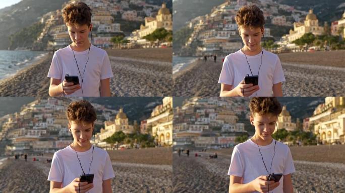 海滩上的小男孩用智能手机。沟通概念。青少年通过手机上的应用程序听音乐和发短信。背景上的古老欧洲意大利