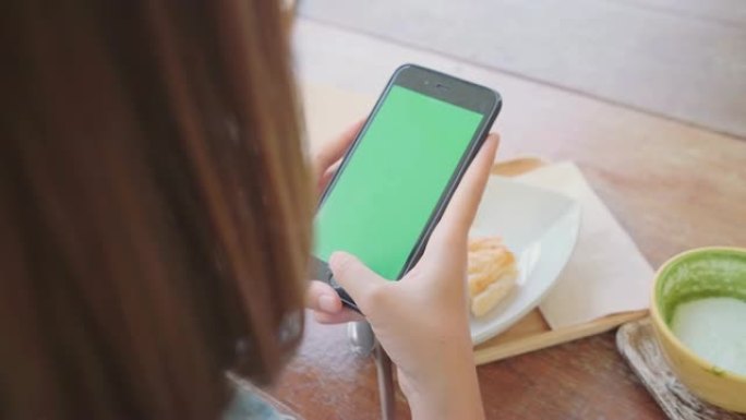 年轻的亚洲妇女使用绿色屏幕的黑色手机设备。亚洲女人拿着智能手机，坐在咖啡馆里滚动页面。色度键。