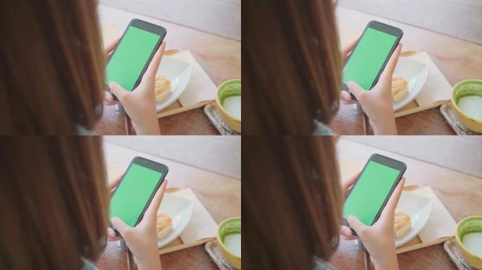 年轻的亚洲妇女使用绿色屏幕的黑色手机设备。亚洲女人拿着智能手机，坐在咖啡馆里滚动页面。色度键。