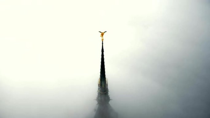 无人机在浓雾上方上升，在神秘的圣米歇尔山城堡尖顶上露出雄伟的金色天使雕像。