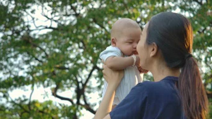 妈妈在公园和婴儿玩耍。年轻的亚洲母亲带着幸福的情感在公园或花园高高举起她的宝贝儿子。慢动作。