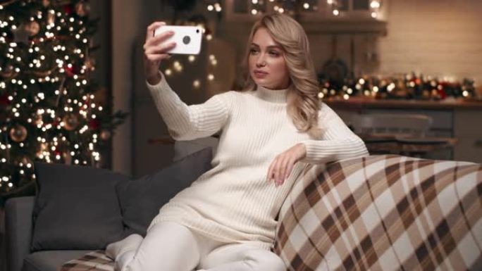 美丽的金发女郎在家中在圣诞树的背景下在客厅制作圣诞节自拍照。
