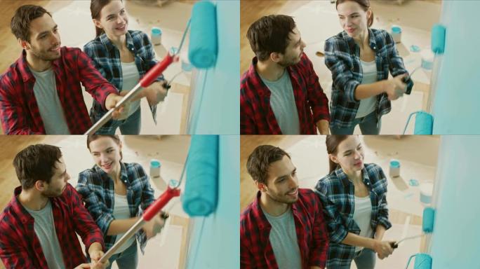 年轻漂亮的夫妇交谈并装饰他们的新公寓。丈夫和妻子正在用蘸有浅蓝色油漆的滚筒粉刷墙壁。他们开心又笑。家