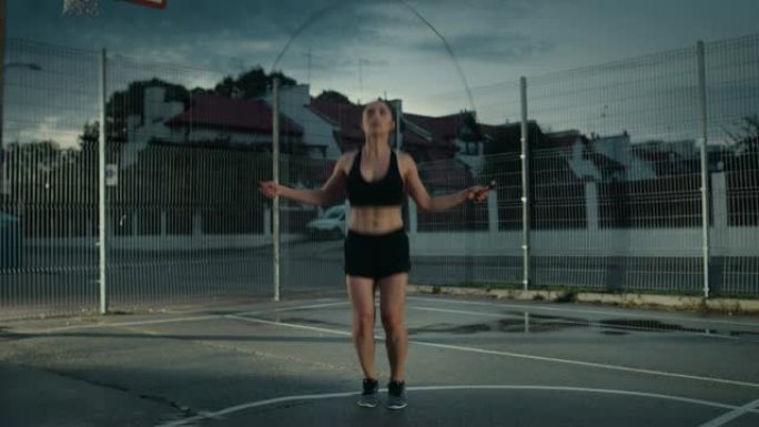 美丽精力充沛的健身女孩跳绳。她正在一个有围栏的室外篮球场里锻炼身体。居民区下雨后的晚间录像。