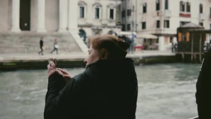 后视图兴奋的活跃的白人老年旅游妇女在威尼斯之旅的贡多拉游览中拍摄智能手机照片。