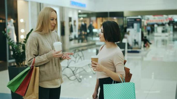 有吸引力的年轻女性在购物中心里聊天，拿着可购买的咖啡和纸袋。人体模型和玻璃墙上的时髦服装是可见的。