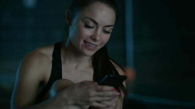 一个穿着黑色运动服的美丽运动健身女孩在晚上使用智能手机时微笑的特写镜头。