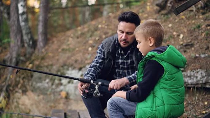 可爱的小男孩正在和爸爸在森林里的池塘里钓鱼，而他的爸爸正在和他交谈，解释如何使用装备。家庭、爱好和自