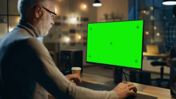 坐在办公桌前的创意中年设计师使用带有绿色模拟屏幕的台式计算机。晚上，两名创意员工在办公室的电脑上工作