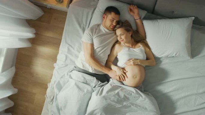 幸福的年轻夫妇在床上拥抱在一起，年轻的女人怀孕了，有爱心的伴侣温柔地抚摸着她的肚子。自上而下的摄像头