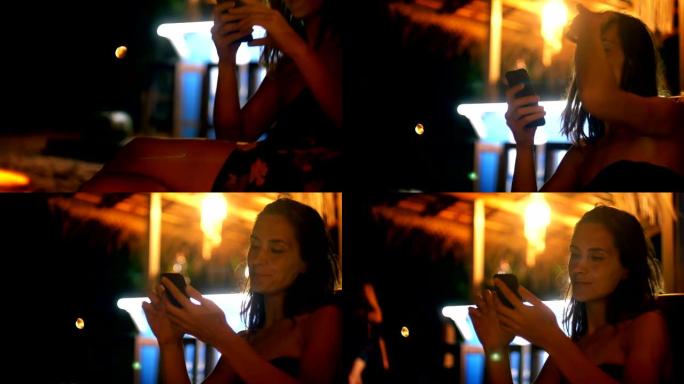 特写镜头倾斜坐在夜间休息室沙滩吧椅上的快乐年轻女子，双腿交叉使用智能手机