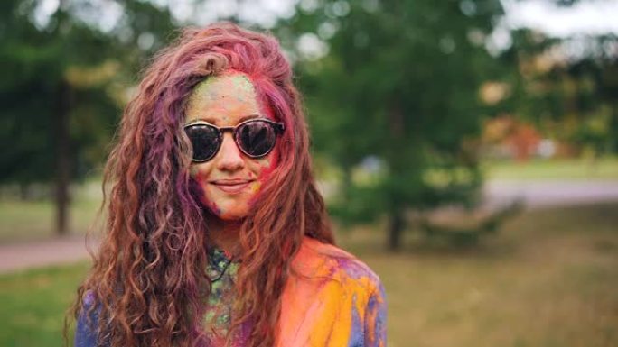 在Holi节上，站在户外，戴着墨镜微笑着，长卷发的快乐女孩的慢动作肖像覆盖着多色油漆粉末。