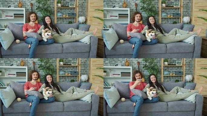 年轻的家庭和柯基犬在家里的沙发上看电视，笑着放松