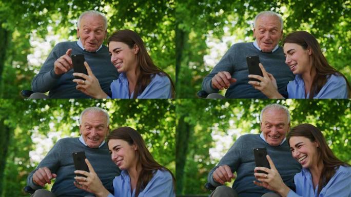 坐在轮椅上的快乐孙女和祖父的慢动作在阳光明媚的日子里，在绿色公园的手机上看他们的照片或在互联网上导航