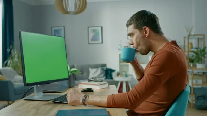 英俊的微笑男人坐在家里的办公桌旁，使用带有绿屏模型的个人计算机。他住在舒适的公寓里，室内有现代客厅。
