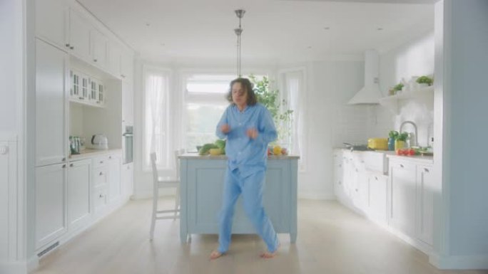 穿着蓝色睡衣的长发在厨房跳舞的疯狂有趣的年轻人。明亮的白色现代厨房区，桌子上有健康的绿色食品。舒适的