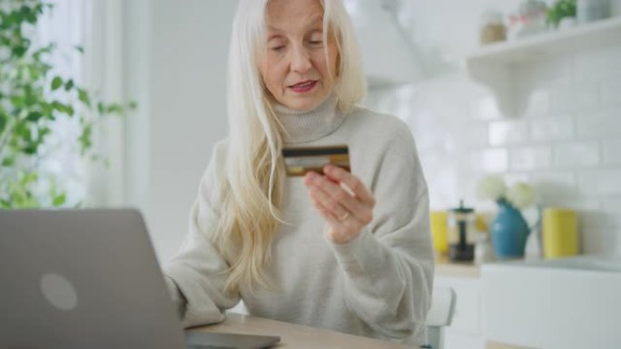 地道的高级女性在家里的厨房里使用笔记本电脑。美丽的白发老年女性养老金领取者使用信用卡在互联网上购物。