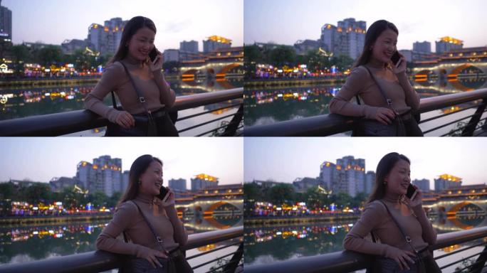 漂亮的亚洲年轻女子戴着墨镜在成都城市大街九岩桥河边散步。美丽的长发女孩在手机上聊天步行旅行在傍晚的中