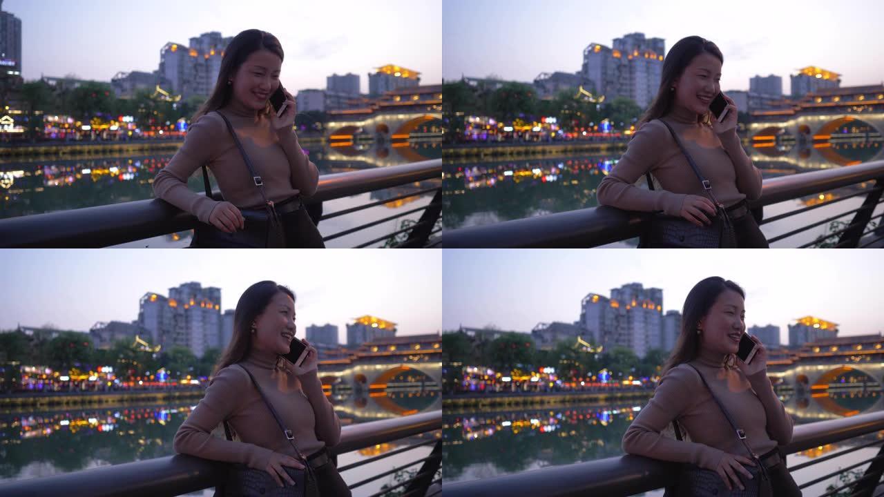 漂亮的亚洲年轻女子戴着墨镜在成都城市大街九岩桥河边散步。美丽的长发女孩在手机上聊天步行旅行在傍晚的中