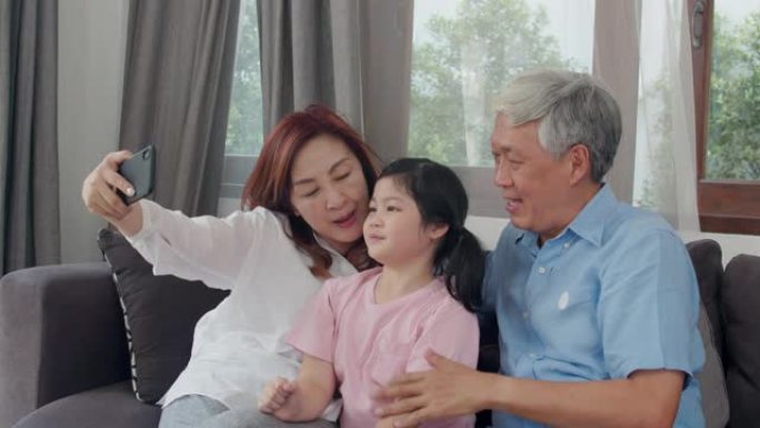 亚洲祖父母在家与孙女自拍。资深中国人，爷爷和奶奶开心地度过家庭时光，用手机放松身心，年轻的女孩孩子躺