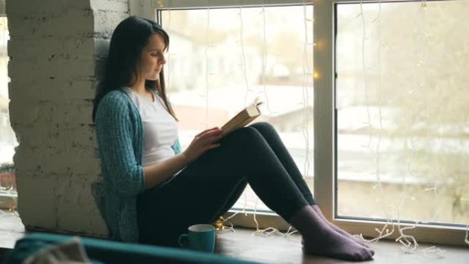 美丽的年轻女子专注于看书，坐在家里的窗台上，然后看着窗外。爱好、舒适和房屋概念。