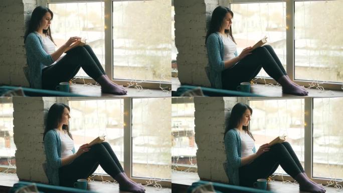 美丽的年轻女子专注于看书，坐在家里的窗台上，然后看着窗外。爱好、舒适和房屋概念。