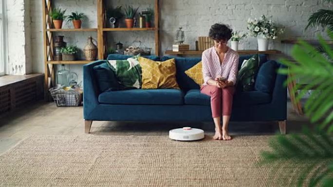 迷人的女人正在打开坐在沙发上的机器人真空吸尘器，然后使用智能手机并在设备清洁房屋时休息。技术和公寓概