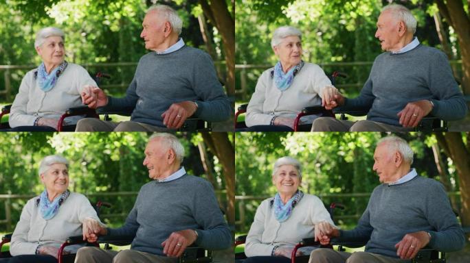 一对老年夫妇的慢动作都坐在轮椅上，彼此深情地牵着手，以示他们永远的爱，尊重和在阳光明媚的日子里在绿色