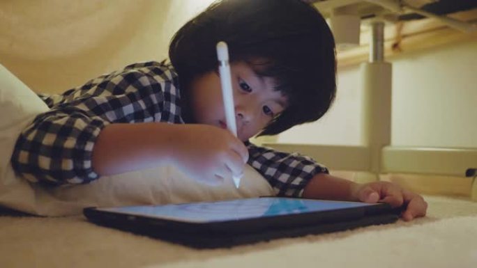 亚洲男孩躺在床上的帐篷下用数字平板电脑做作业