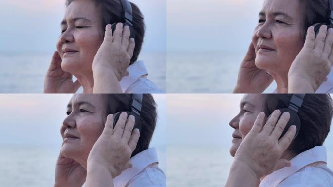 日落时在美丽的大海前听音乐时戴着耳机的快乐亚洲老年妇女的肖像。假期