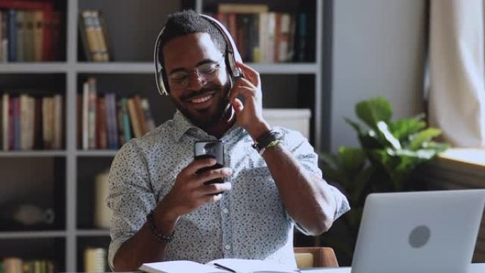 戴着耳机微笑的非洲男子在办公桌前听手机音乐